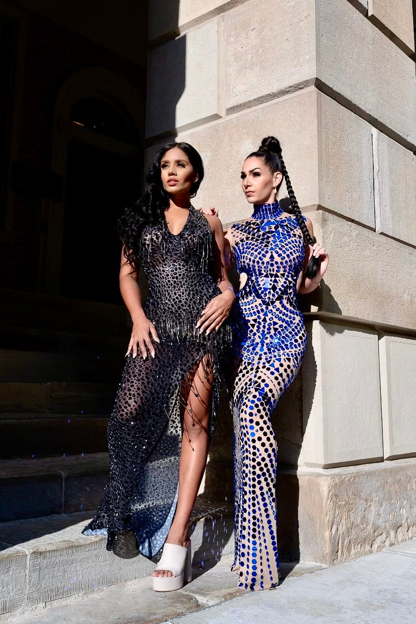 Two women in elegant dresses standing against a stone building.